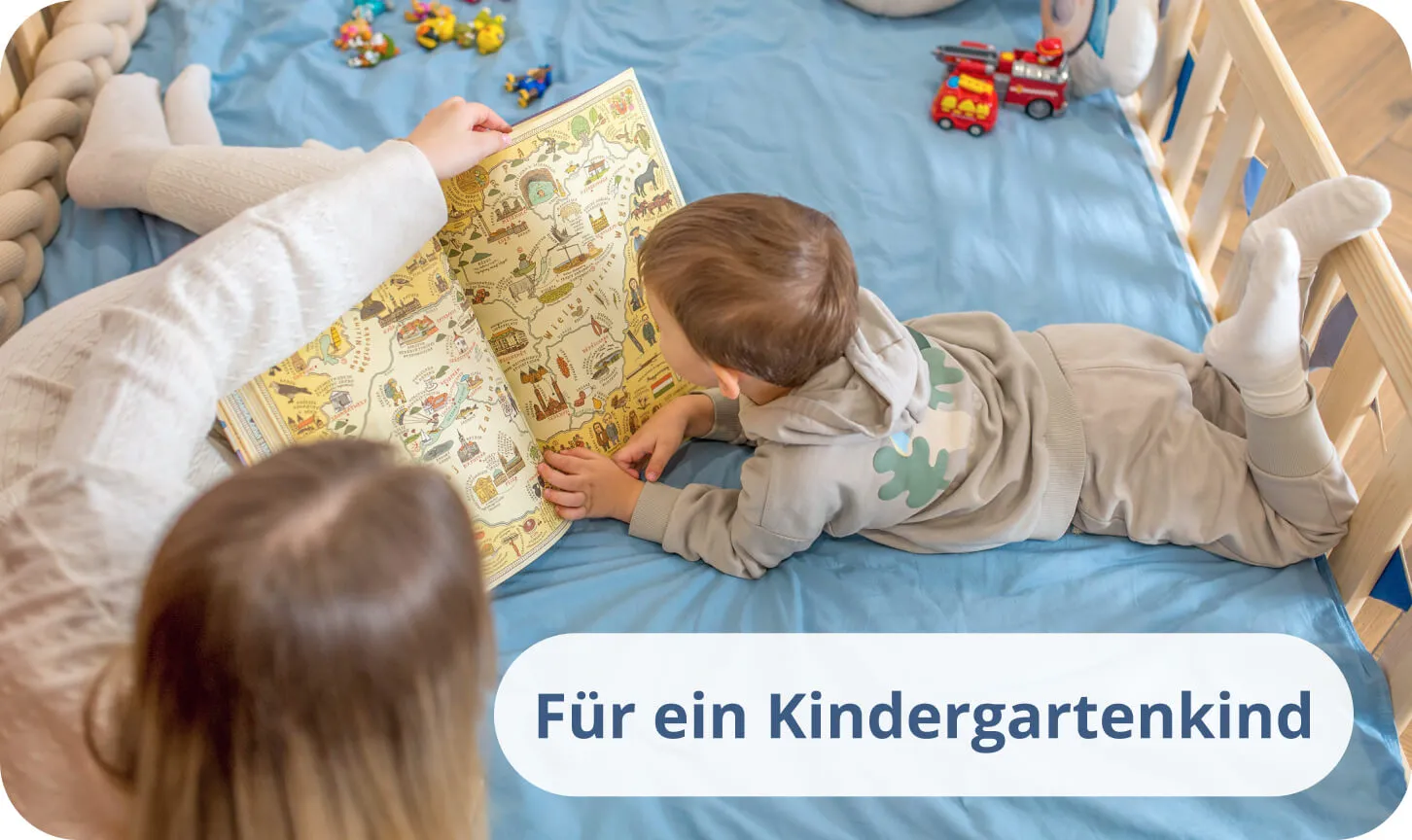 Kleiner Junge mit seiner Mutter liest ein buntes Buch auf blauer Bettwäsche in einem Kiefernholzbett.